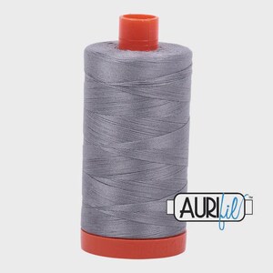 Puede incluir: Una bobina de hilo gris con tapa y base naranja. El hilo está bien enrollado alrededor de la bobina. La marca "AURIfil" está impresa en el lateral.