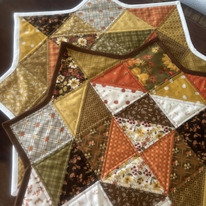 May include: A star-shaped quilted table runner featuring a patchwork design of various fabrics in shades of orange, yellow, brown, and cream. The fabric patterns include floral prints, plaid, and small pumpkin motifs, finished with a white border.