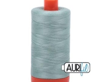 May include: A spool of light blue thread with a red base and top. The thread is tightly wound, and the spool has the Aurifil logo on the side. This thread is likely used for sewing, quilting, or embroidery projects.