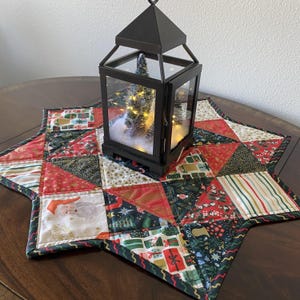 Könnte beinhalten: Eine schwarze Metalllaterne mit einem kleinen Weihnachtsbaum im Inneren steht auf einem sternförmigen, gesteppten Tischläufer. Der Läufer hat ein Patchwork-Design mit rot-, grün- und goldfarbenen Weihnachtsmotiven. Die Laterne hat einen Griff oben.