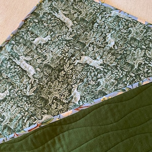 May include: A quilted blanket featuring a forest green base with wavy stitching. The top section has a dark green floral pattern with white rabbits. The edge is trimmed with a light blue floral fabric.