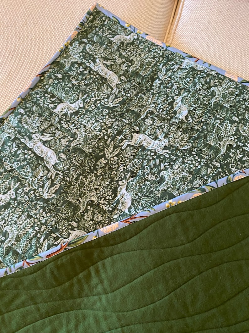 May include: A quilted blanket featuring a forest green base with wavy stitching. The top section has a dark green floral pattern with white rabbits. The edge is trimmed with a light blue floral fabric.