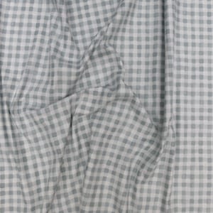 May include: A close-up of a fabric with a gingham pattern. The fabric is a light blue-gray color with a white grid pattern. The fabric is draped, showing folds and shadows.