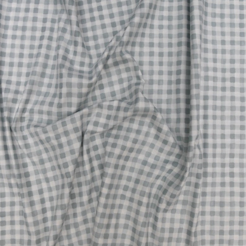 May include: A close-up of a fabric with a gingham pattern. The fabric is a light blue-gray color with a white grid pattern. The fabric is draped, showing folds and shadows.