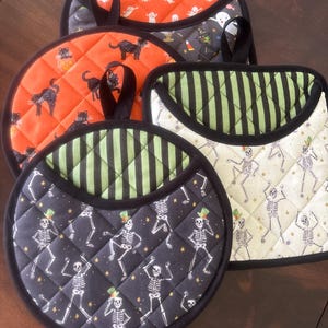 Puede incluir: Un conjunto de tres agarradores acolchados con estampados de Halloween. Uno es naranja con gatos negros, otro blanco con esqueletos bailando y otro negro con esqueletos bailando, todos con ribetes y lazos para colgar en color negro.