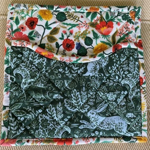 May include: Quilted pot holder with a floral and woodland design. The top section features a white background with red poppies, yellow flowers, and green leaves. The bottom section has a dark green background with white squirrels and rabbits.