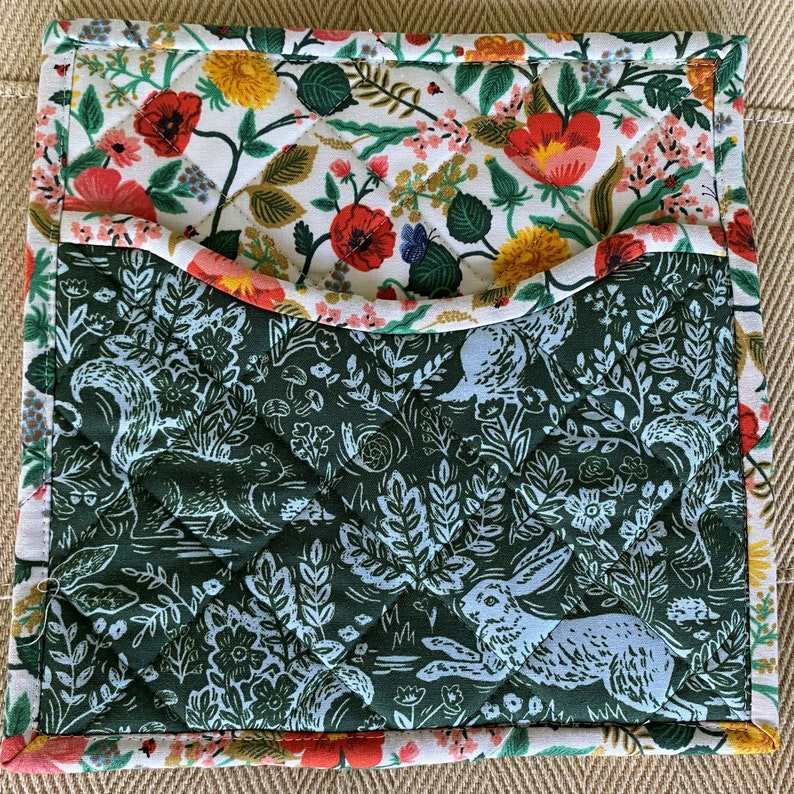 May include: Quilted pot holder with a floral and woodland design. The top section features a white background with red poppies, yellow flowers, and green leaves. The bottom section has a dark green background with white squirrels and rabbits.