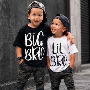 Pode incluir: Duas crianças com roupas a combinar, consistindo em calças com estampa de camuflagem e ténis vermelhos. Uma criança usa uma t-shirt preta com o texto branco BIG BRO e um boné. A outra usa uma t-shirt branca com o texto preto LIL BRO.