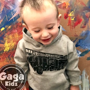 May include: A young child wearing a grey hoodie with a black and white illustration of a fire engine. The hoodie has the text "Gaga Kidz" on the front. The child is standing in front of a colourful painted wall.
