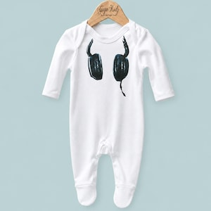 May include: A white long-sleeve baby bodysuit featuring a black graphic print of headphones on the chest. The garment is displayed on a wooden hanger labeled Gaga Kidz, with a round neckline and integrated footed bottoms for full coverage.