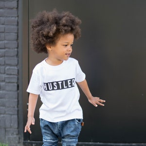 May include: A young boy with curly brown hair is wearing a white t-shirt with the word "Hustler" printed in black on it. He is also wearing blue jeans and black sneakers.