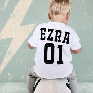 Könnte beinhalten: Ein weißes kurzärmeliges T-Shirt mit dem Namen EZRA in fetten schwarzen Blockbuchstaben auf dem oberen Rücken, wobei die Zahl 01 in einer größeren, passenden schwarzen Schriftart direkt unter dem Namen auf dem Rücken aufgedruckt ist.