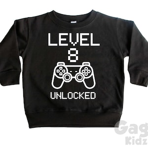 May include: Black long-sleeve sweatshirt with white text that reads "LEVEL 8 UNLOCKED" and a white graphic of a video game controller.