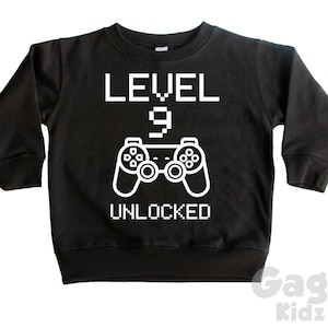 Op de afbeelding: Zwart longsleeve sweatshirt met witte tekst die "LEVEL 9 UNLOCKED" zegt en een witte afbeelding van een videogamecontroller.