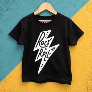 Könnte beinhalten: Ein schwarzes kurzärmeliges T-Shirt mit einer großen weißen Blitzgrafik auf der Vorderseite. Im Inneren des Blitzes steht der Text Rock and Roll in einer stilisierten Schrift, ergänzt durch kleine Sterne und Striche. Das T-Shirt hängt an einem Kleiderbügel.