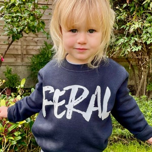 May include: A black sweatshirt with the word "FERAL" written in white paint across the front. The sweatshirt is being worn by a young child.