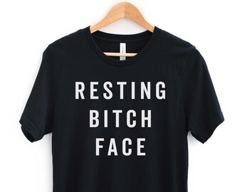 May include: Black t-shirt with the phrase "RESTING BITCH FACE" printed in white block letters. The shirt is hanging on a wooden hanger against a white background. The Gaga Kidz logo is in the bottom right corner.