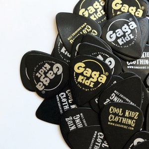 Puede incluir: Un montón de púas de guitarra negras con letras doradas y plateadas. Las púas presentan el texto "Gaga Kidz" y "Cool Kidz Clothing" con la dirección del sitio web "www.gagakidz.com".