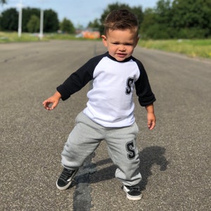 May include: A young child wearing a white and black baseball tee with a large black letter 'S' on the front and matching grey sweatpants with a black letter 'S' on the leg. The child is standing on a paved road.