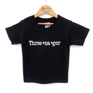 K&ouml;nnte beinhalten: Schwarzes T-Shirt mit wei&szlig;em Text "Three-na-ger" auf einem Holzb&uuml;gel.