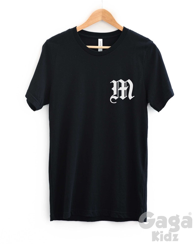 K&ouml;nnte beinhalten: Schwarzes T-Shirt mit einem wei&szlig;en "M" in gotischer Schrift auf der Vorderseite.