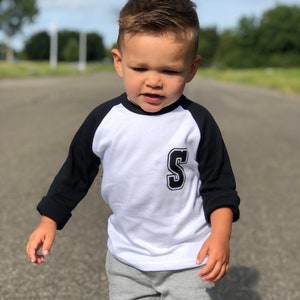 May include: A young child wearing a white and black baseball tee with a black 'S' on the front and a grey pair of sweatpants with a black 'S' on the leg.