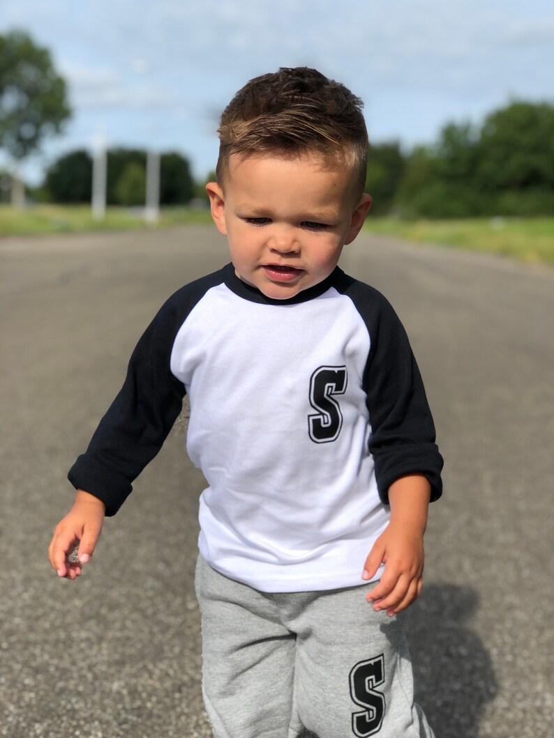 May include: A young child wearing a white and black baseball tee with a black 'S' on the front and a grey pair of sweatpants with a black 'S' on the leg.