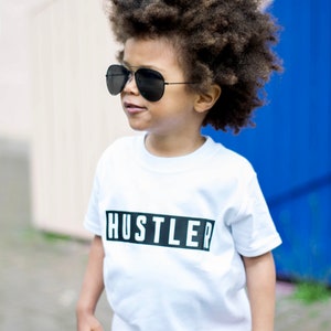 May include: A young person wearing a white t-shirt with the word "Hustler" printed in black on a black rectangle. The person is wearing black sunglasses and orange shorts. The person has curly brown hair.
