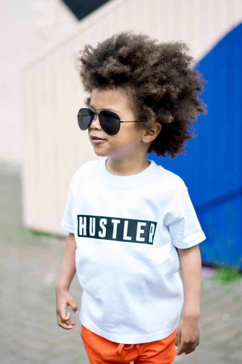 May include: A young person wearing a white t-shirt with the word "Hustler" printed in black on a black rectangle. The person is wearing black sunglasses and orange shorts. The person has curly brown hair.