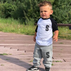 May include: A young child wearing a white and black baseball shirt with the letter 'S' on the front and grey sweatpants with the letter 'S' on the leg. The child is wearing black trainers.