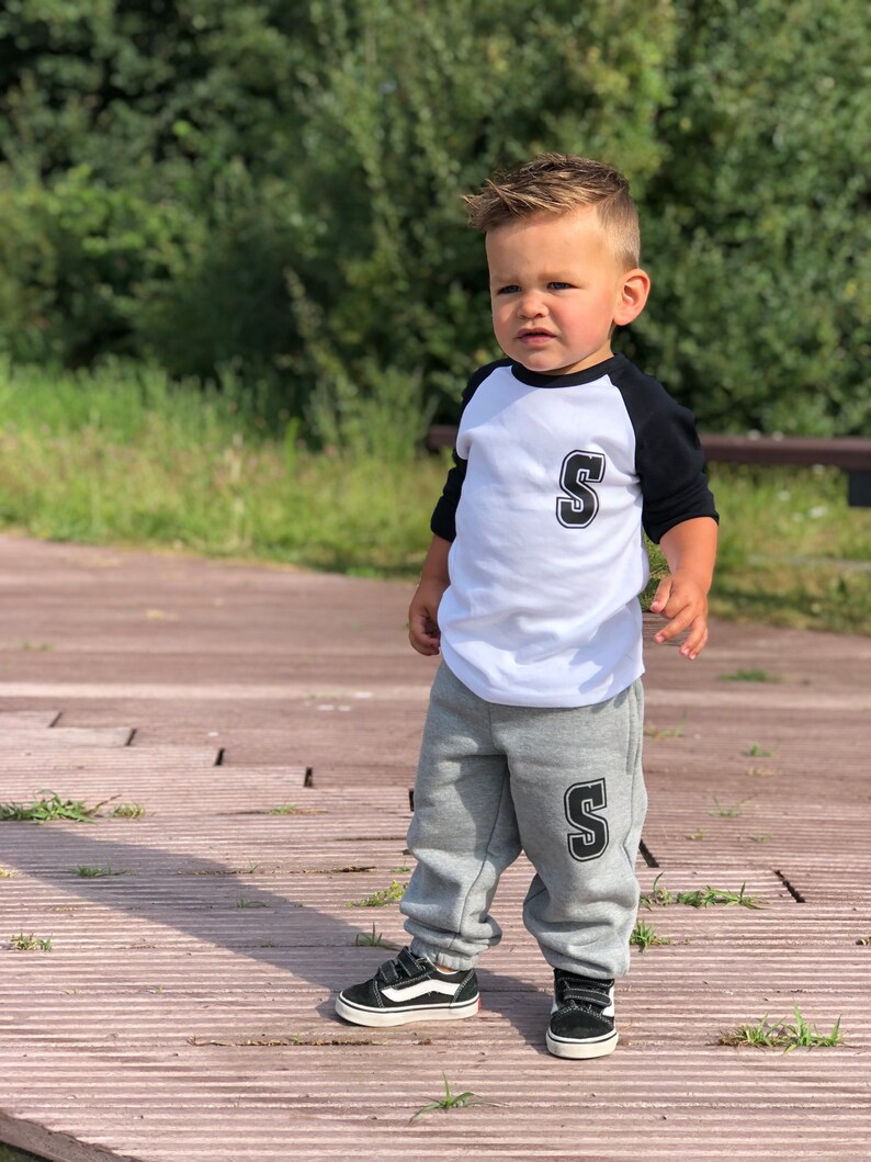 May include: A young child wearing a white and black baseball shirt with the letter 'S' on the front and grey sweatpants with the letter 'S' on the leg. The child is wearing black trainers.