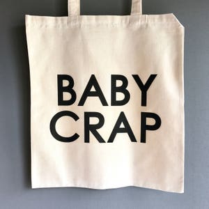 Pode incluir: Uma bolsa de lona branca com texto preto que diz "BABY CRAP".