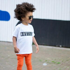 May include: A young boy wearing a white t-shirt with the word "HUSTLE" printed on it in black. He is also wearing orange pants and black sneakers. He is standing in front of a white and black wall.