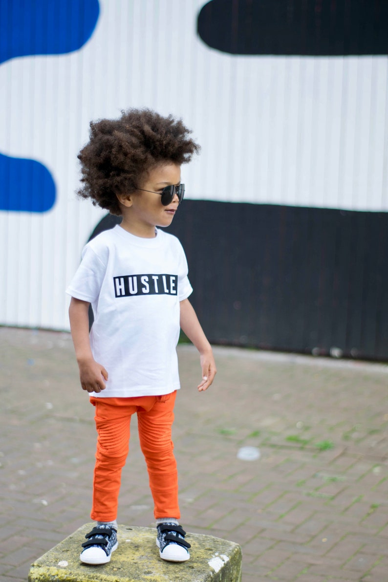 May include: A young boy wearing a white t-shirt with the word "HUSTLE" printed on it in black. He is also wearing orange pants and black sneakers. He is standing in front of a white and black wall.