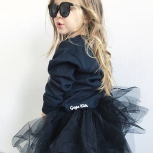 Pu&ograve; includere: Una bambina che indossa una felpa nera con la scritta "Gaga Kids" sul davanti e una gonna di tulle nera. Indossa occhiali da sole neri.