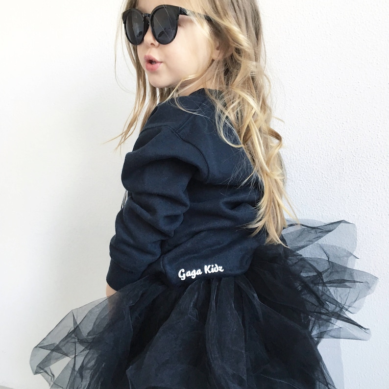 Pu&ograve; includere: Una bambina che indossa una felpa nera con la scritta "Gaga Kids" sul davanti e una gonna di tulle nera. Indossa occhiali da sole neri.