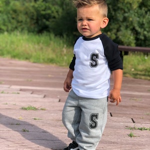 May include: A young boy wearing a white and black baseball shirt with a black letter 'S' on the front and grey sweatpants with a black letter 'S' on the left leg. He is wearing black and white trainers.