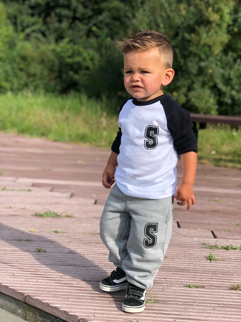 May include: A young boy wearing a white and black baseball shirt with a black letter 'S' on the front and grey sweatpants with a black letter 'S' on the left leg. He is wearing black and white trainers.