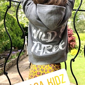 May include: A gray hooded sweatshirt featuring the white text WILD & THREE printed on the back, paired with yellow and black leopard print leggings. A white rectangular label displays the yellow text @GAGA_KIDZ across the lower portion.