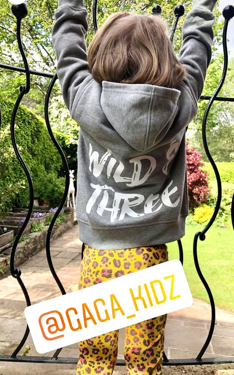 May include: A gray hooded sweatshirt featuring the white text WILD & THREE printed on the back, paired with yellow and black leopard print leggings. A white rectangular label displays the yellow text @GAGA_KIDZ across the lower portion.