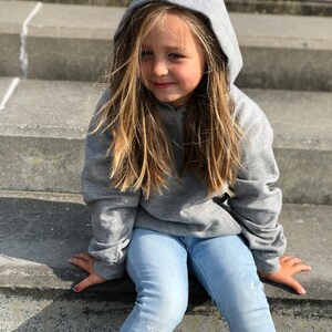 May include: A heather gray long-sleeved hooded sweatshirt with a front kangaroo pocket, paired with light blue denim jeans featuring subtle distressed detailing on the thighs. The hoodie is worn with the hood pulled up over the head.