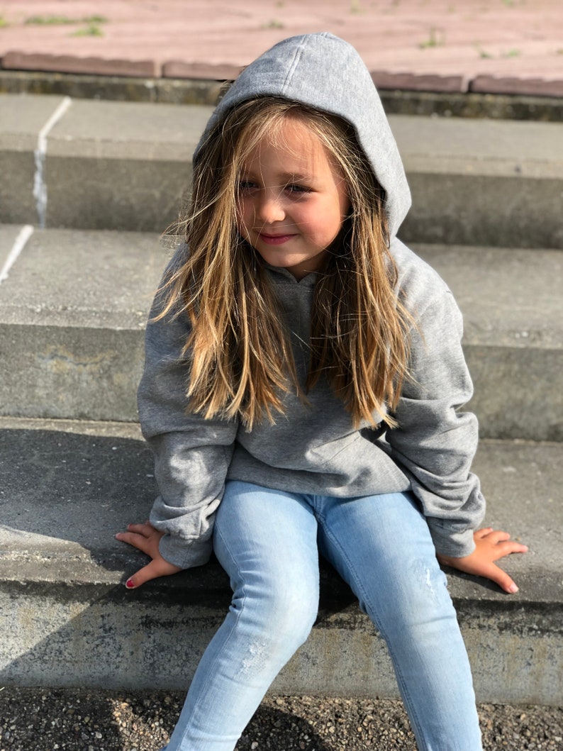 May include: A heather gray long-sleeved hooded sweatshirt with a front kangaroo pocket, paired with light blue denim jeans featuring subtle distressed detailing on the thighs. The hoodie is worn with the hood pulled up over the head.