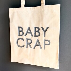 Pode incluir: Uma bolsa de lona branca com texto preto que diz "BABY CRAP".