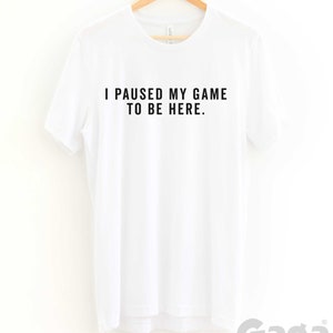 K&ouml;nnte beinhalten: Wei&szlig;es T-Shirt mit schwarzem Text, der "Ich habe mein Spiel pausiert, um hier zu sein." lautet.