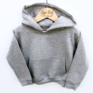 May include: A heather gray long-sleeve hooded sweatshirt featuring a front kangaroo pocket, ribbed cuffs, and a ribbed hem. The hoodie is displayed on a wooden hanger with the text Gaga Kidz Rock the High Chair printed in brown.