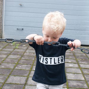May include: A young boy wearing a black t-shirt with the word "HUSTLER" printed in white on it. He is holding a metal chain in his hands and looking down at it.