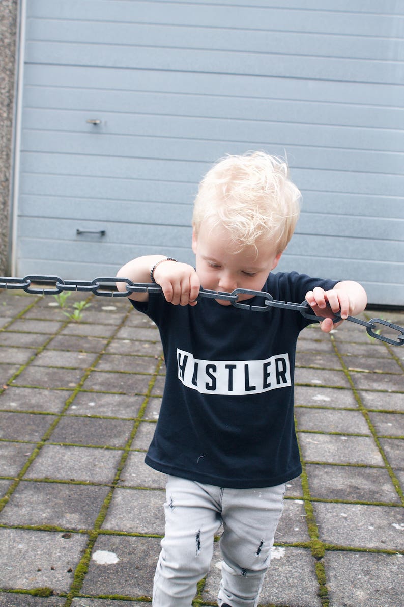 May include: A young boy wearing a black t-shirt with the word "HUSTLER" printed in white on it. He is holding a metal chain in his hands and looking down at it.