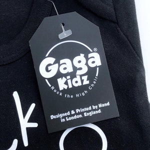Op de afbeelding: Zwart label met witte tekst die "Gaga Kidz - Rock the High Chair" en "Designed & Printed by Hand in London, England." zegt.