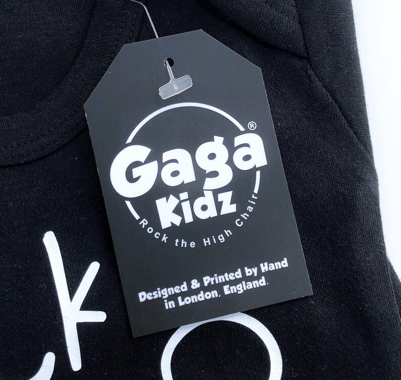 Op de afbeelding: Zwart label met witte tekst die "Gaga Kidz - Rock the High Chair" en "Designed & Printed by Hand in London, England." zegt.