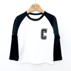 May include: White long-sleeved baseball tee with black sleeves and a black felt letter "C" on the front. The shirt is hanging on a wooden hanger with the text "Gaga Kidz Rock the High Chart" on it.